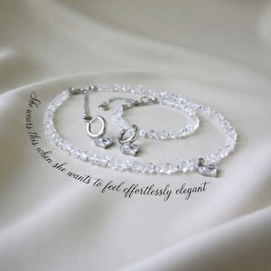 May include: A clear crystal necklace and matching earrings set. The necklace and earrings are made of small, faceted crystals and silver-toned metal. The text in the image reads: "She wears this when she wants to feel effortlessly elegant."