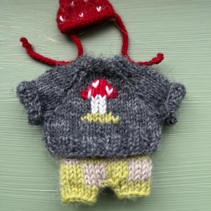 May include: A hand-knitted outfit consisting of a dark gray sweater with a central red and white mushroom motif, a pair of light green and beige checkered shorts, and a matching red pointed hat with white accents and long red tie strings.