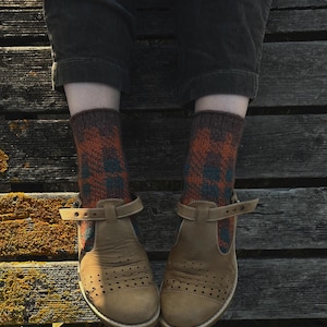 Pode incluir: Um par de sapatos de couro bege com tira em T, biqueiras perfuradas e fivelas laterais ajustáveis, usados com meias de malha até o meio da panturrilha com padrão xadrez castanho-escuro e laranja, combinados com calças de veludo cinzentas.