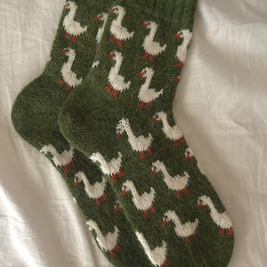 Puede incluir: Un par de calcetines verdes de punto con un patrón repetido de gansos blancos con patas rojas. Los calcetines están hechos de un hilo texturizado y se muestran sobre una superficie blanca. Un accesorio acogedor y caprichoso.