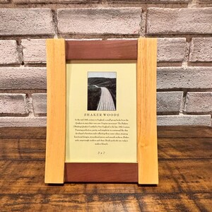 May include: A wooden picture frame with a landscape photo. The frame has a light wood color on the sides and a darker brown wood at the top and bottom. The photo is of a road. The text "SHAKER WOODS" is visible.