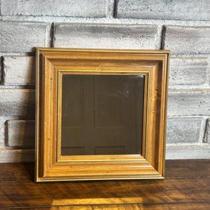 May include: Square wooden framed mirror with a gold trim. The mirror is set against a gray brick wall and rests on a wooden surface. The frame has a natural wood grain finish.