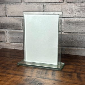 May include: A vertical rectangular glass picture frame featuring a clear glass border and a central white display area. The frame is supported by a thick, rectangular glass base, providing a stable foundation for the upright display structure.