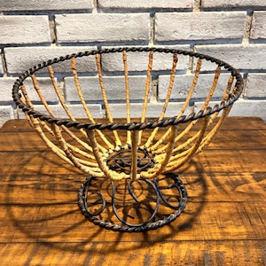 May include: A decorative fruit basket with a woven, light brown interior and a dark, twisted metal frame. The basket has a round base and a flared rim, and stands approximately 10 inches tall.