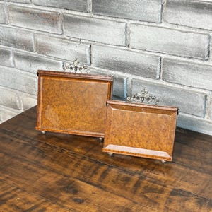 May include: Two decorative wooden trays with ornate silver-colored accents. The trays have a warm, brown wood grain pattern and are rectangular with small feet. The trays are displayed on a dark wooden surface against a brick wall.