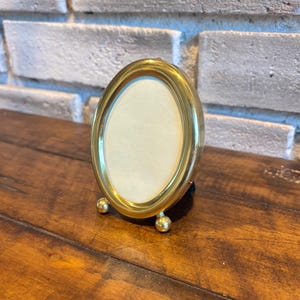 Könnte beinhalten: Ein kleiner, ovaler Bilderrahmen in Goldfarbe. Der Rahmen hat ein schlichtes Design mit glatter, polierter Oberfläche. Er steht auf drei kleinen, runden Füßen. Der Rahmen ist leer, mit einer cremefarbenen Rückseite.