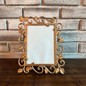 May include: A gold-colored, rectangular picture frame with an ornate, leaf-and-vine design. The frame has a white interior and is displayed on a dark wooden surface. The background is a light-colored brick wall.