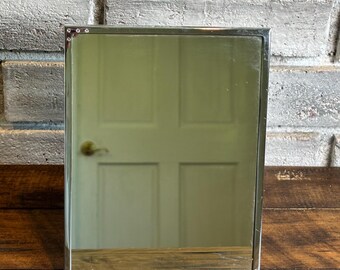 May include: A rectangular tabletop mirror featuring a polished silver-tone metal frame. The frame is accented with small, clear, circular decorative studs at each of the four corners, providing a subtle detail to the clean, metallic border.