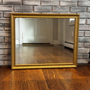 May include: A rectangular wall mirror featuring a wide, ornate gold-toned frame with a textured, speckled finish. The mirror glass is clear and reflective, showing a view of an interior space with wooden flooring and a white door in the reflection.