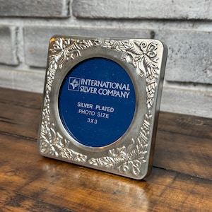 May include: A square silver-plated photo frame with a circular center opening and embossed floral corner details. The blue insert features the text International Silver Company, Silver Plated, Photo Size 3x3 inches in white lettering.