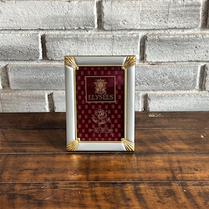 May include: A rectangular white picture frame with gold-colored accents at the corners. The frame holds a burgundy-colored insert with the word "ELYSEES" in gold lettering. The frame is displayed on a wooden surface.