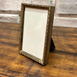 May include: A small, rectangular gold-colored picture frame with a decorative, textured border. The frame has a white interior and a black easel back, perfect for displaying a photograph or artwork. The frame is standing on a wooden surface.