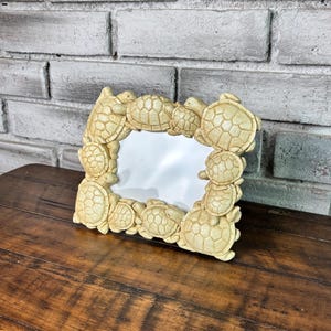 May include: A rectangular photo frame with a cream-colored finish, adorned with numerous turtle figurines. The frame is made of a composite material and has a white interior. The frame is placed on a dark wooden surface.