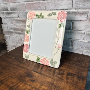 May include: Cream-colored rectangular picture frame decorated with pink roses and green leaves. The frame is made of ceramic or a similar material and is displayed on a dark wooden surface.