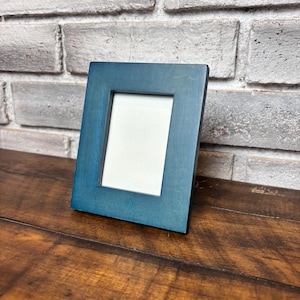 May include: A rectangular picture frame with a smooth, matte teal blue finish. The frame features a wide, flat border surrounding a centered, blank white rectangular insert, designed to hold a photograph or artwork in a vertical orientation.