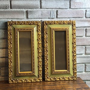 May include: A pair of rectangular mirrors featuring ornate gold-toned frames with intricate scrollwork detailing and a sage green inner border. The mirrors have a vertical orientation and a reflective glass center, showcasing a classic design aesthetic.