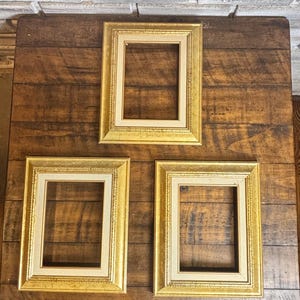 May include: Three rectangular picture frames with gold-colored borders and off-white matting are arranged on a dark wooden surface. The frames are empty, ready for photos or artwork. The frames have a classic, ornate design.