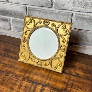 May include: A square picture frame with a metallic gold finish featuring a circular center opening. The frame has a beaded inner border and raised scrollwork patterns in each corner, creating a textured surface around the central display area.