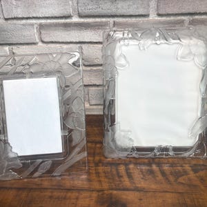 May include: Two clear glass picture frames featuring a frosted, raised relief design of daffodils and stems. The frames are rectangular with smooth edges and a central white rectangular insert for displaying photographs or artwork.