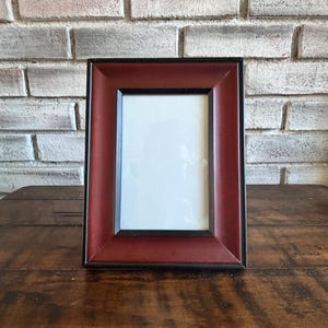 May include: A rectangular picture frame with a dark red border and a black inner frame. The frame is empty, with a white space for a photo. The frame is sitting on a wooden surface, with a brick wall in the background.