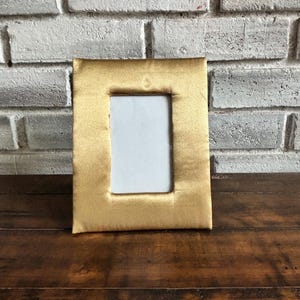 Puede incluir: Un marco de fotos rectangular dorado con interior blanco. El marco está hecho de un material suave y texturizado. Se encuentra sobre una pared de ladrillos blancos y una superficie de madera oscura.