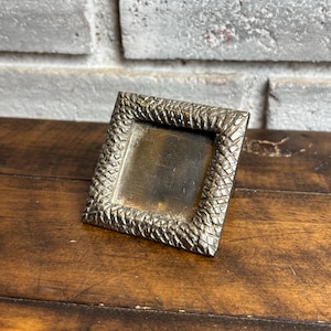 May include: A small square picture frame with a metallic finish and a textured, rope-like border pattern. The center of the frame is a smooth, dark, reflective surface. The frame is angled slightly, showing its depth and detailed outer edge.
