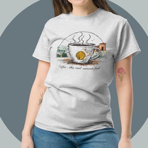 May include: A white ceramic mug with a black handle and the text "But First, Coffee" in a bold, black font. The mug is set against a blurred background, suggesting a cozy setting.