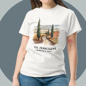 Op de afbeelding: Wit T-shirt met een aquarel illustratie van een weg met cipressen en de tekst "VIA FRANCIGENA - Walking to Rome". Het ontwerp is gecentreerd op het shirt.