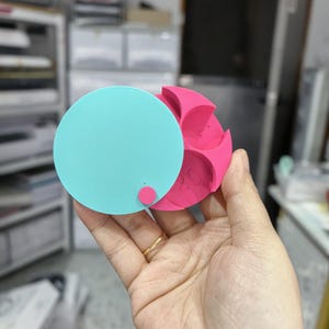May include: A two-part, 3D-printed object. The top is a light blue circle with a small pink peg. The bottom is a pink, multi-lobed shape with the number "20" visible. The object is held in a hand.