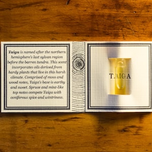 Puede incluir: Un pequeño frasco de vidrio transparente con un líquido amarillo en su interior. El frasco está etiquetado con "TAIGA" y se encuentra en una caja blanca con una descripción del aroma. La caja está sobre una superficie de madera.