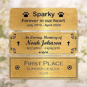 May include: Three rectangular gold-colored plaques with black text. The top plaque reads "Sparky" with paw prints and dates. The middle plaque says "In Loving Memory of Noah Johnson" with dates and crosses. The bottom plaque says "FIRST PLACE SUMMER LEAGUE 2025" with soccer balls.