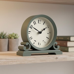 May include: A sage green, round tabletop clock with a cream-colored face and black numerals and hands. The clock has a decorative base with scroll-like accents. The clock is displayed on a light-colored wooden shelf.