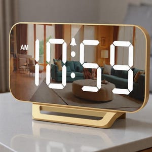 May include: A digital alarm clock with a gold-colored frame and base displays the time as 10:59 AM in bright white digits. The clock's mirrored face reflects a living room scene. The clock is set on a light-colored surface.