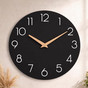 May include: A round, black wall clock with white numerals and light brown hands. The clock face has a minimalist design, with the numbers 1 through 12 clearly displayed. The hands are made of wood. A modern home decor item.