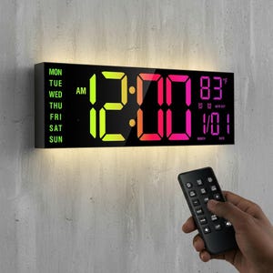 May include: Black digital wall clock displaying the time as 12:00 AM, with the temperature at 83°F and the date 1/01. The clock has a black frame and a remote control is in the foreground.