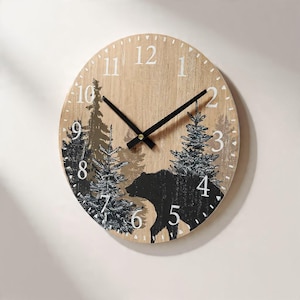 May include: Round wooden wall clock with a forest scene featuring a black bear silhouette and evergreen trees. The clock face has white numerals and black hands. The clock is mounted on a light-colored wall.