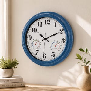 May include: A round wall clock with a distressed blue frame and a white face featuring large black Arabic numerals. Two smaller circular gauges are positioned at the bottom, displaying humidity and temperature readings in Fahrenheit.