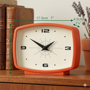 May include: A retro-style orange clock with a cream-colored face. The clock measures 7 inches wide and 5 1/4 inches tall. Black hands and numerals mark the time. The clock has a mid-century modern design.