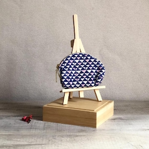 Pode incluir: Uma bolsa redonda com padrão geométrico azul marinho e branco com zíper dourado, exibida em um pequeno cavalete de madeira. A bolsa apresenta um design de triângulo repetido com detalhes em rosa.