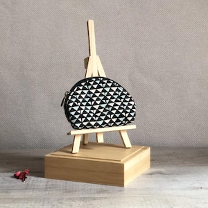 Pode incluir: Uma pequena bolsa redonda com um padrão geométrico em preto e branco e fecho de correr. A bolsa é exibida em um cavalete de madeira em miniatura e um bloco de madeira. O fundo é cinza neutro.