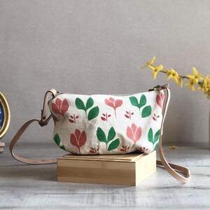Può includere: Una borsa a tracolla bianca con un motivo floreale di tulipani rossi e foglie verdi. La borsa ha una tracolla in pelle marrone e poggia su una scatola di legno. Una stampa botanica incorniciata e un vaso di fiori gialli sono sullo sfondo.