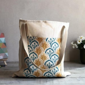 May include: A cream-coloured canvas tote bag featuring a large front pocket with a repeating pattern of teal and mustard yellow stylised leaves and floral motifs. The bag has two sturdy, matching cream-coloured fabric handles attached at the top.