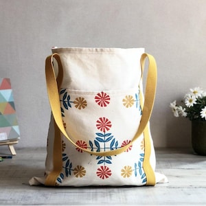 May include: A cream-coloured canvas tote bag featuring a repeating floral pattern in red, blue, and yellow. The bag has two mustard yellow shoulder straps and a large exterior pocket on the front. The design consists of stylised flowers and leaves.