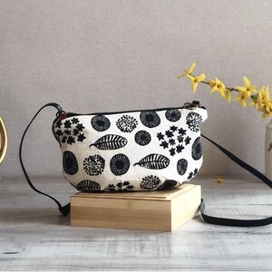 Può includere: Una borsa a tracolla bianca con una stampa botanica nera di foglie e motivi floreali circolari. La borsa è dotata di una chiusura a zip nera e di una sottile tracolla nera. Il tessuto ha un aspetto testurizzato con un motivo grafico ripetuto.
