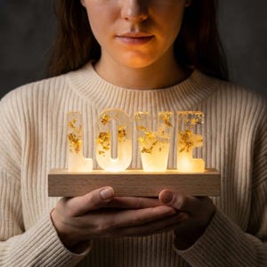 Könnte beinhalten: Ein leuchtendes "LOVE" Schild mit Goldblatt-Akzenten, auf einem Holzsockel. Die Buchstaben sind klar und beleuchtet, was eine warme Atmosphäre schafft. Ein dekoratives Element für die Inneneinrichtung.