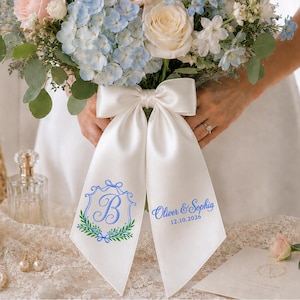 Op de afbeelding: Een wit satijnen lint gestrikt om een boeket van blauwe en witte bloemen. Het lint is voorzien van blauw borduurwerk met een decoratieve letter B in een wapenschild en de tekst Oliver & Sophia met de datum 12.10.2026 in blauw cursief schrift.