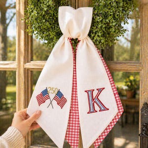 Puede incluir: Colgador de puerta de tela color crema con ribete de cuadros rojos y detalles bordados. Presenta banderas americanas cruzadas con el número 250 y la letra "K" en rojo y azul. Una corona verde es visible en el fondo.