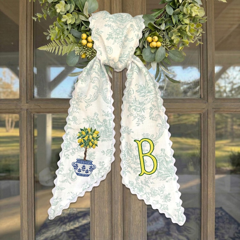 May include: A decorative fabric door hanger featuring a light blue and white toile pattern with scalloped edges. The left side displays a potted lemon tree illustration, while the right side features a large yellow letter B. It is attached to a green wreath.