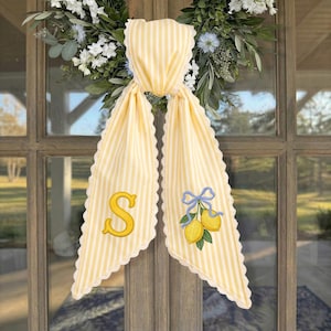May include: A yellow and white striped fabric door hanger with scalloped edges. One side features a large embroidered yellow letter S, while the other side displays an embroidered pair of yellow lemons tied with a light blue ribbon bow.