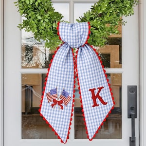 Puede incluir: Bufanda de puerta a cuadros azules y blancos con bordes festoneados rojos, una letra "K" bordada y dos banderas americanas. La bufanda cuelga de una puerta blanca con una corona verde.